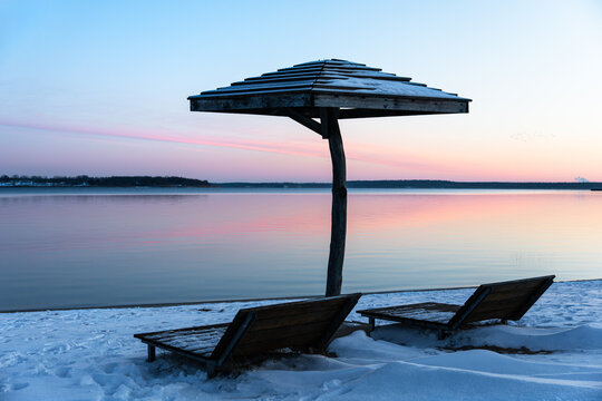 Sunshade with two deck chairs on the snow-covered beach at Goitzsche Lake, winter sunset with pastel sky, serene lakeshore landscape in Bitterfeld.