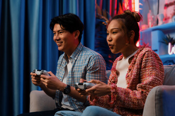 Sitting sofa of happy smiling couple playing video game control by joystick fronted snack and drinks competing challenge level with funny at modern neon blub light comfy living room at home. Infobahn.