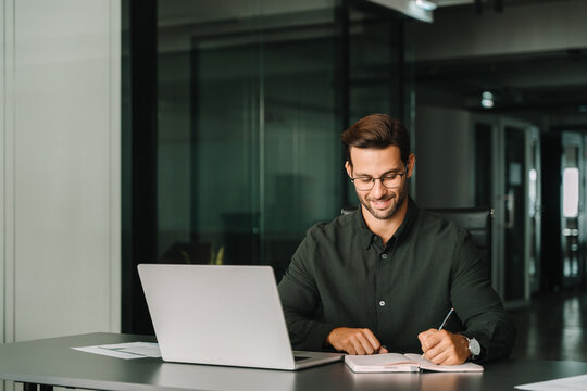 Smiling young man student using laptop computer for business studying watching online webinar e-learning training meeting writing notes. European businessman entrepreneur working in office, copy space