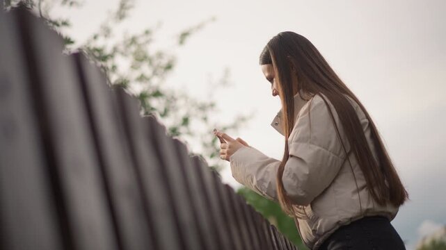 woman checking phone near fence, golden sky backlight with soft highlights, long braids cascading, relaxed posture leaning on rail, casual jacket, scrolling messages, soft bokeh foliage, outdoor calm,
