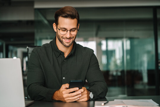 Smiling mature latin Hispanic business manager ceo using cell phone mobile app, laptop. European young student man businessman holding smartphone sitting in office working online on gadget, copy space - Powered by Adobe