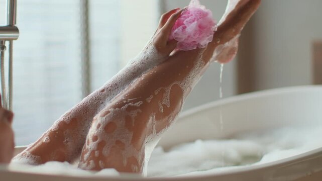 A woman relaxes in a bathtub filled with bubbles. She washes her leg with a bath sponge while water runs. Sunlight streams through a window, creating a warm atmosphere.