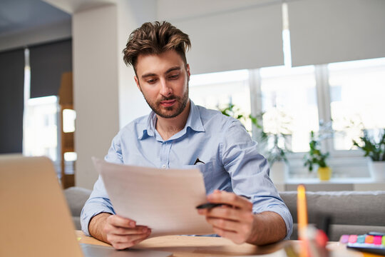 Serious male entrepreneur reviewing business documents and analyzing paperwork in home office.