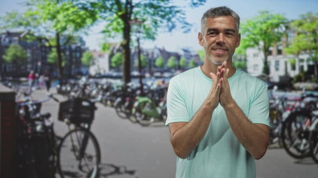 Man with clasped hands beside bicycles on a street waterfront, smiling with stubble and gray hair; quiet gratitude reflection.