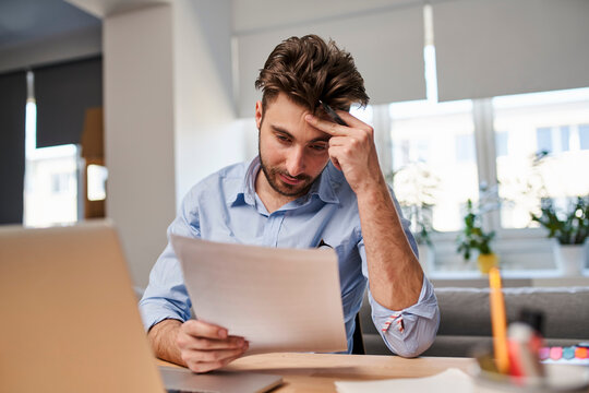Stressed man reviewing documents at home office desk. Financial problems and paperwork stress concept.