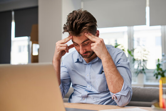Stressed man suffering from headache and burnout at home office. Overwhelmed male worker touching head in frustration.