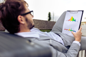 Young businessman lying on a bench outdoors, reviewing a paper report.