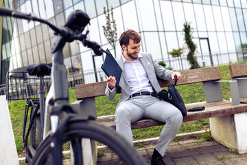 Young businessman placing his tablet into a bag while standing in a city environment with a bicycle.