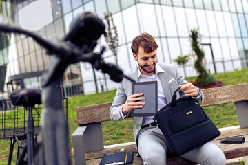 Young businessman placing his tablet into a bag while standing in a city environment with a bicycle.