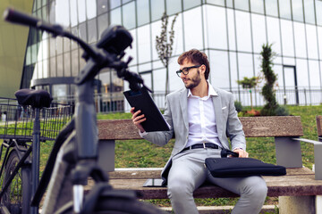 Smiling young businessman holding a tablet while standing next to his bicycle in a city environment.