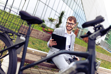 Smiling young businessman holding a tablet while standing next to his bicycle in a city environment.