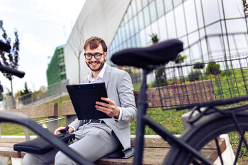 Smiling young businessman holding a tablet while standing next to his bicycle in a city environment.