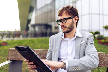 Young businessman sitting on a bench outdoors, carefully reviewing a paper report.