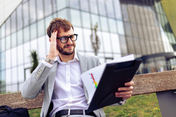Young businessman sitting on a bench outdoors, holding his head while reviewing a paper report.