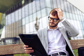 Young businessman sitting on a bench outdoors, holding his head while reviewing a paper report.