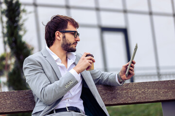 Young businessman sitting on a bench in front of a modern office building, drinking coffee and using a tablet.