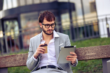 Young businessman sitting on a bench in front of a modern office building, drinking coffee and using a tablet.