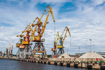Fototapeta premium Industrial harbor cranes lined along a port quay, used for cargo handling and maritime logistics under a cloudy sky