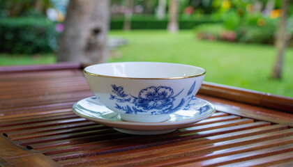 A delicate blue and white teacup with gold trim sits on a wooden table in a serene garden setting.
