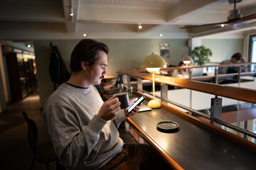 Relaxed man customer drinking coffee and checking news on smartphone in modern bookstore cafe space. Smart focused guy having lunch break in cozy library with co-working space using cellphone.