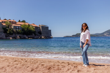 A woman in blue jeans and a white shirt stands on a sandy beach by the sea on a sunny day. Relaxed summer vibes, coastal scenery, clear water, travel, relaxation, and tourism