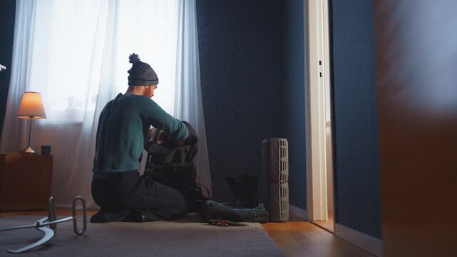 Man packing winter gear and survival tools into backpack at home, indoor preparation scene