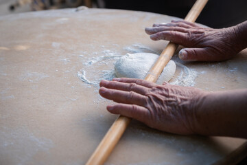 Elderly hands actively working, rolling fresh white dough on a light wooden surface dusted with flour, demonstrating a traditional baking method and artisanal craft