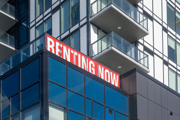 Low angle view of a modern apartment building with a red and white Renting Now sign on a glass balcony. Contemporary residential high-rise architecture featuring blue glass windows, metal cladding