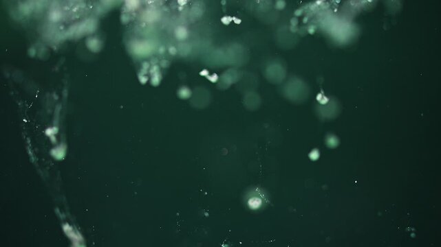 Macro shot of green bubbles and particles floating in liquid with a dark green background and soft bokeh depth of field. Slow motion