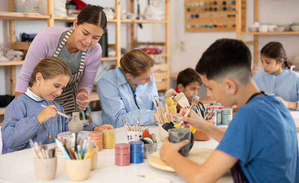 Children and teenagers sit at a table and paint ceramics at the workshop. Teacher shows how to paint a clay product after firing