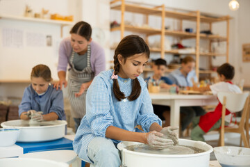 Interested preteen girl making clay pot using potter's wheel in art studio together with others