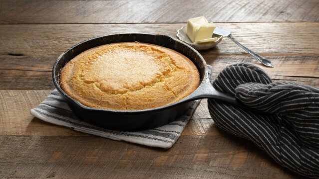Golden brown, freshly baked, buttermilk cornbread in a cast iron skillet with a black and white striped oven mitt on a wooden table.