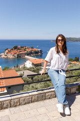 Young woman in casual clothes sitting on the railing of the observation deck overlooking the sea and the island in the background, summer vacation, travel and tourism concept, space for text