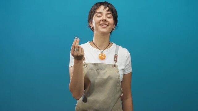 Woman jeweler holding a ring between fingers in studio wearing apron and rings, short hair, eyes closed and smiling; contentment.