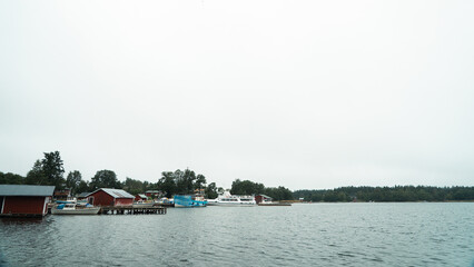 Fototapeta premium Quiet harbor in the Finnish archipelago with boats and red wooden buildings, calm sea landscape and overcast sky with wide copy space