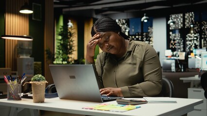 African american woman realizing she works alone late at night in the office, pushing limits despite clear signs of fatigue and tension. Workplace challenges and discouragement. Camera A.