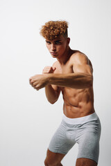 Fototapeta premium Athletic young man with curly hair practicing boxing punch in gray shorts against white background. Muscular fit male training for fitness and combat sport workout.