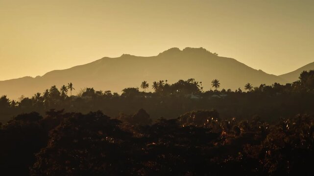 Lombok, Indonesia, Mount Rinjani and the surrounding Senaru village mountains silhouetted against a golden sunrise sky, with tropical palm trees and lush forest below. Aerial rise up panorama
