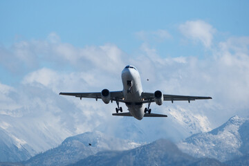 Obraz premium Takeoff with Stunning Mountain Backdrop. White airplane taking off against snow-capped mountains, clouds and a blue sky.