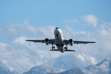 Naklejka premium Takeoff with Stunning Mountain Backdrop. White airplane taking off against snow-capped mountains, clouds and a blue sky.