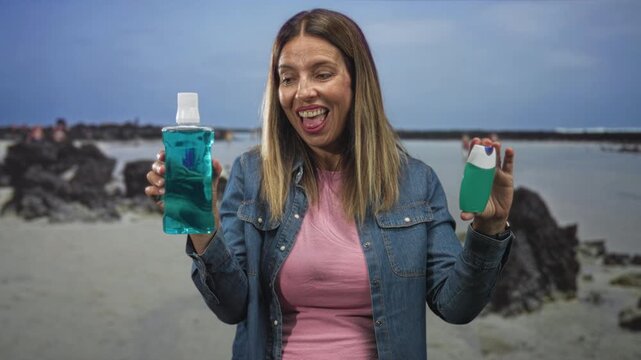 Middle aged woman holds mouthwash bottle and travel toothpaste, smiles showing teeth and leans forward at beach with rocks and tide pools visible; oral hygiene cheerful.