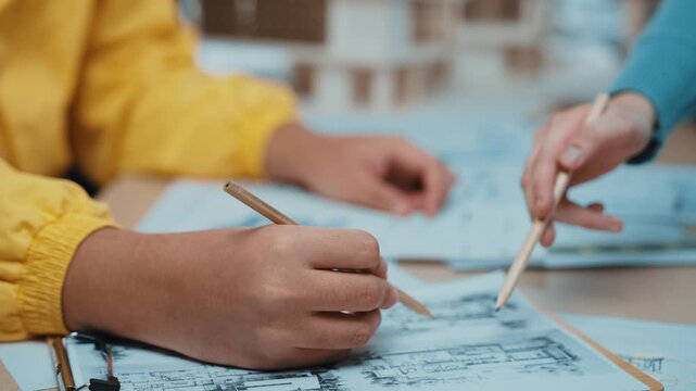 A young person hand is captured in a close-up while sketching architectural plans on a table. Nearby tools and blueprints enhance the creative workspace atmosphere. SACTR