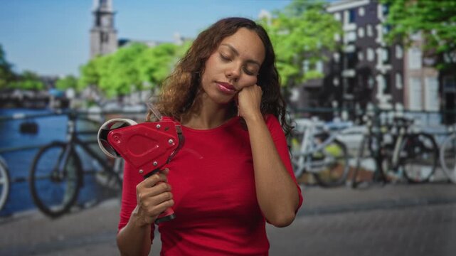Woman holding red tape dispenser and hand on cheek on street in amsterdam near canal with bicycles and brick buildings; tired solitude.