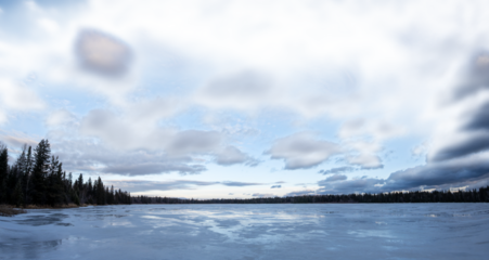 View across an ice covered lake at a spruce forest with a partly transparent sky
