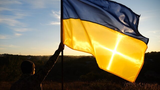 Young soldier of ukrainian army lifted blue-yellow banner in honor of the victory against russian aggression at sunset. Male military in uniform raised a waving flag of Ukraine at countryside