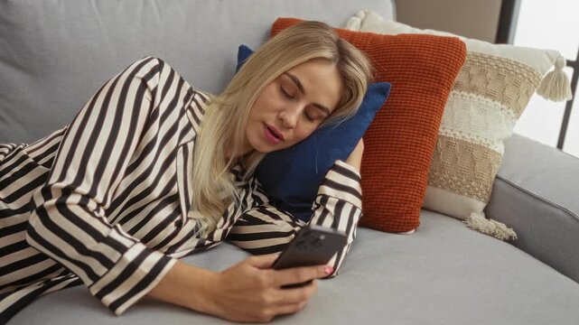 Woman tapping smartphone screen while reclining on a sofa with patterned pillows in a modern apartment; leisure comfort serenity relaxation.