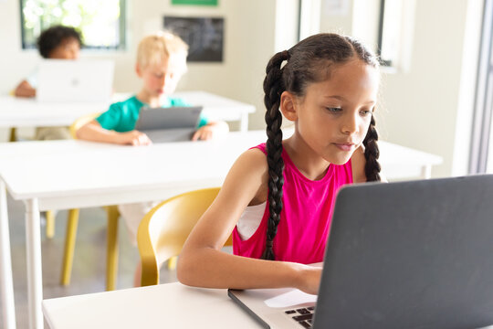 Diverse teenage classmates sitting in classroom, girl working on laptop with tablet nearby