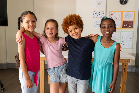 Four youths smiling and posing shoulder-to-shoulder in classroom with wooden table and drawings