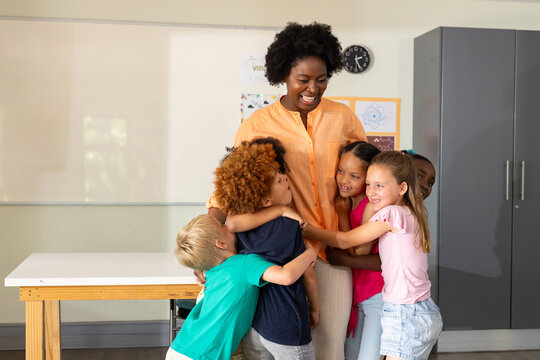 African American teacher in light orange blouse smiling and hugging kids in classroom with clock