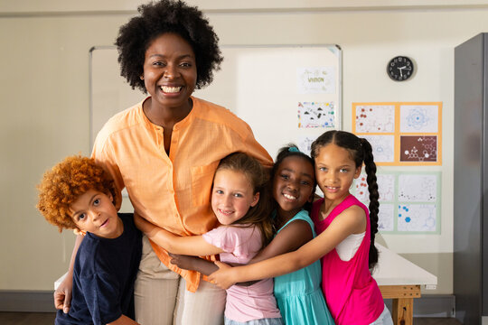 African American teacher smiling while children hugging her in classroom with whiteboard and clock